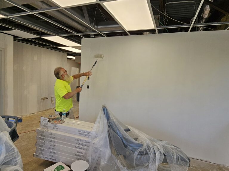 A person wearing a bright yellow shirt is painting a white wall with a roller in a room under renovation. The floor is covered with supplies, and chairs are wrapped in plastic. The ceiling has exposed panels and lighting fixtures.