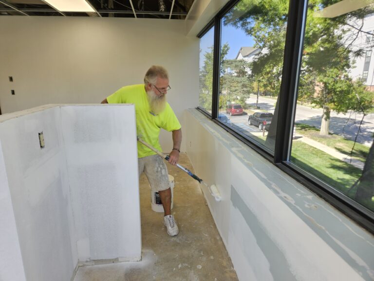 A person with a beard, wearing a bright yellow shirt, is using a paint roller to paint a wall near a large window. The room has unfinished walls and a partially open ceiling. Outside, trees and parked cars are visible.