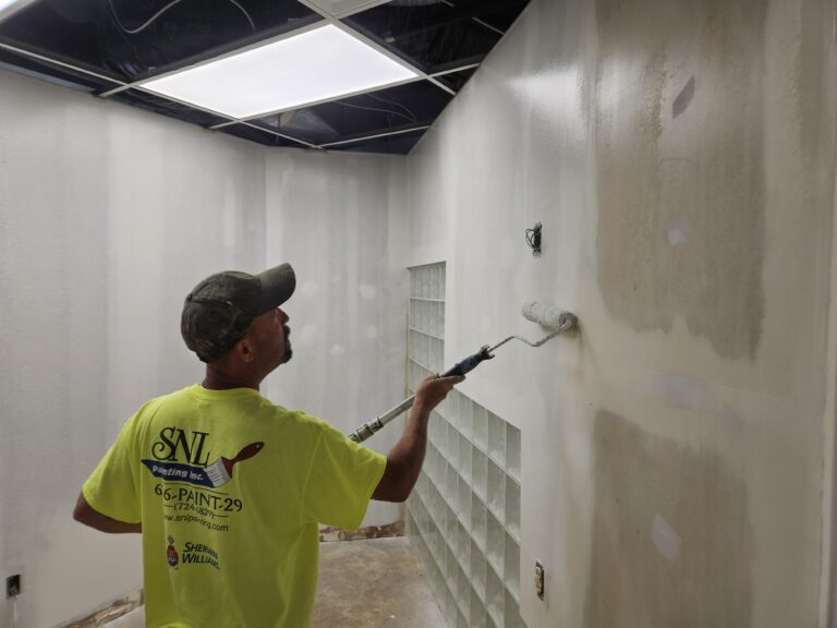 A person wearing a bright yellow shirt and a cap uses a roller brush to paint the wall of a white room. The ceiling tiles are partially missing, revealing the structure above. There is a glass block window in the wall.