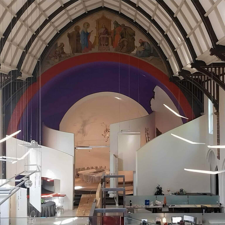 A modern office space is set inside a repurposed church with high arched ceilings and a mural above. Partitioned white walls create work areas below the painted arch, blending contemporary and historical elements.